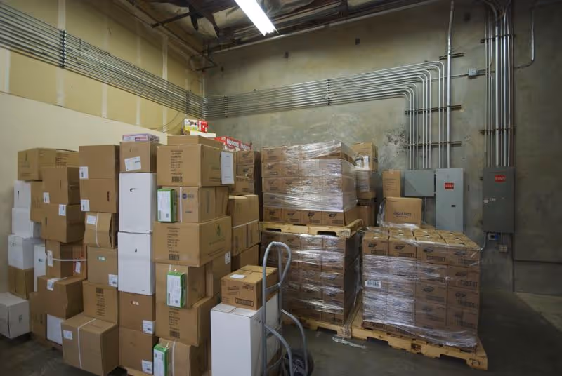 A storage room filled with stacked cardboard boxes on pallets and exposed pipes.