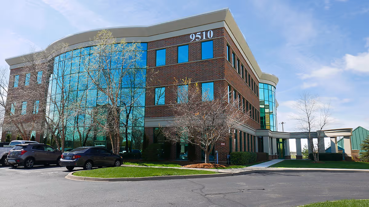 Modern brick building with glass windows, entrance, and surrounding parking lot under blue sky.