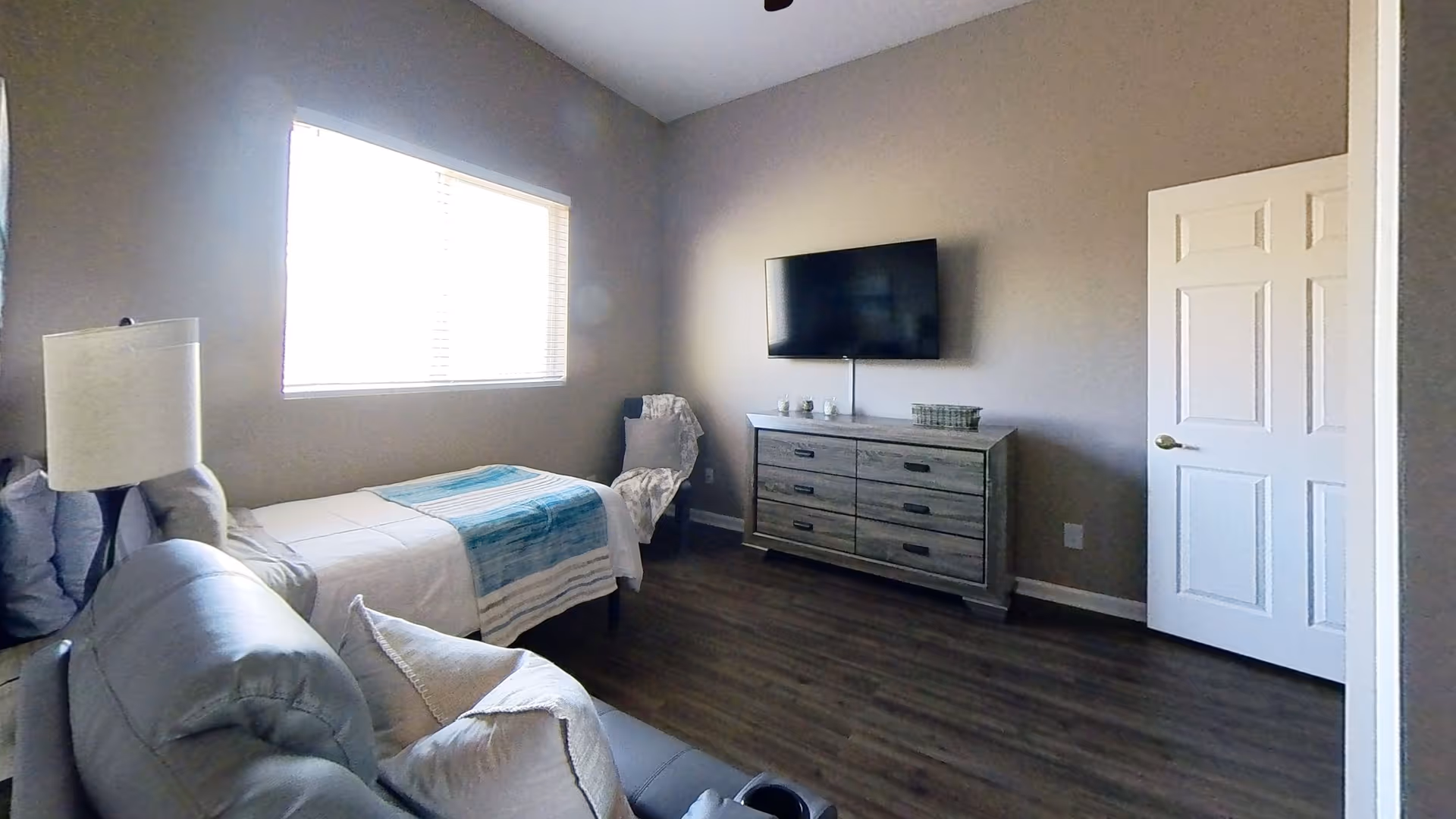 Bedroom in a senior living facility with a bed, dresser, TV, and window.