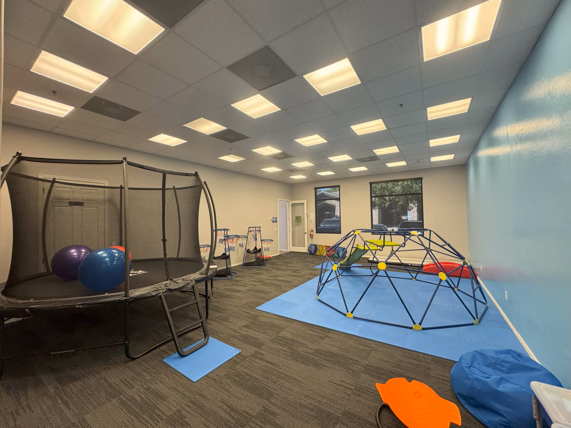 An indoor therapy playroom with a trampoline, climbing dome, mats, and colorful equipment.