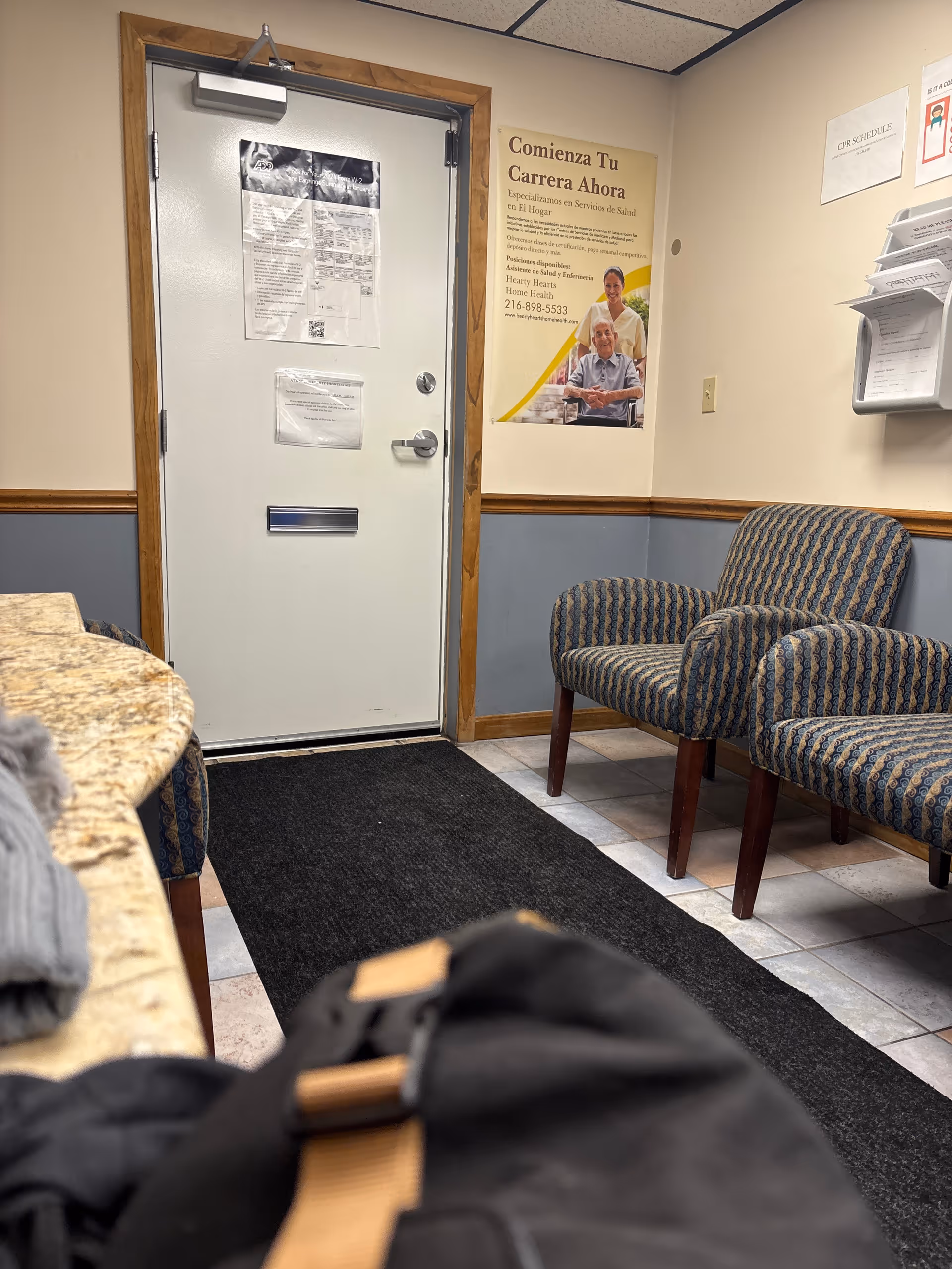Interior waiting area with patterned chairs, a door, posters, and a rug.