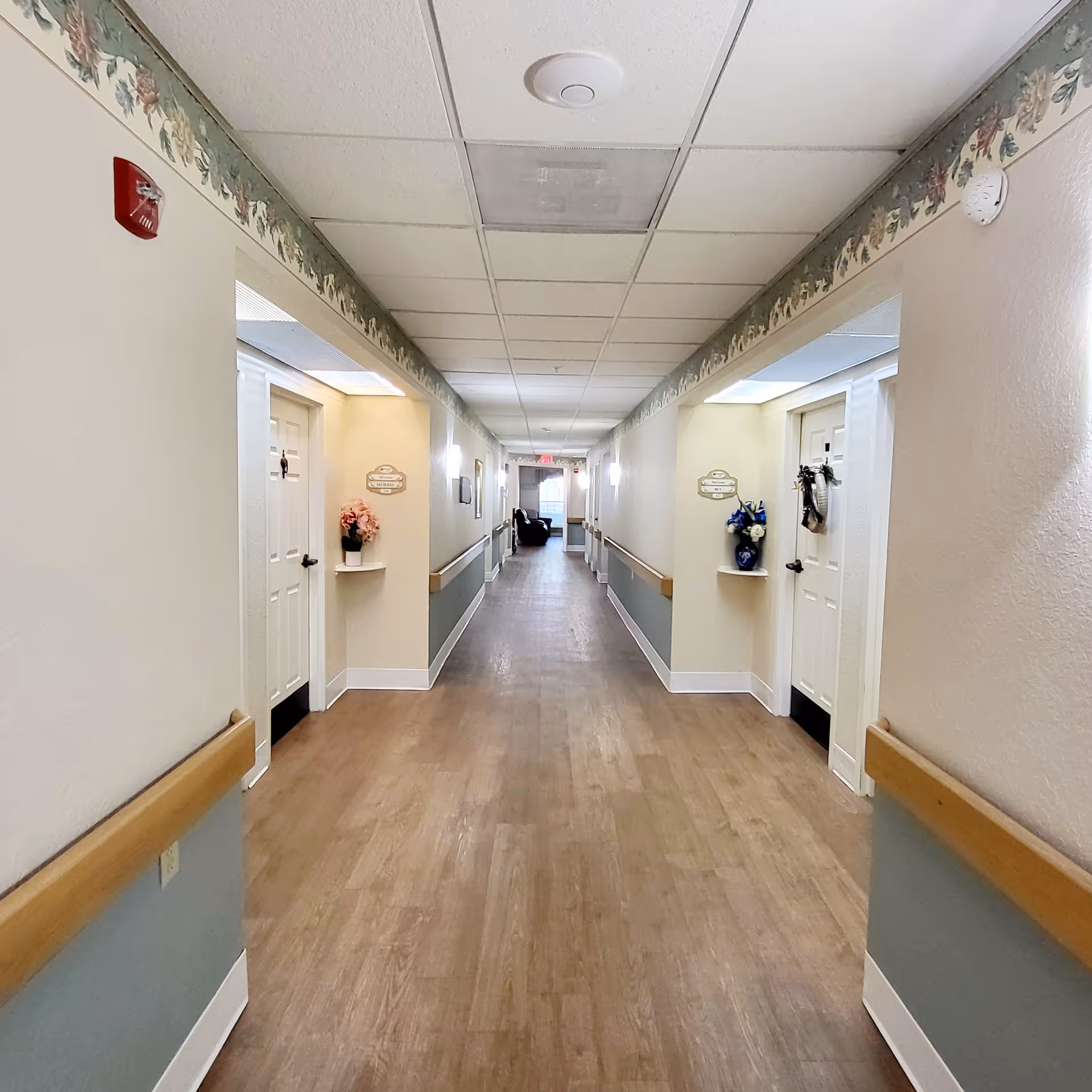 Long, well-lit hallway in a senior living facility with doors and floral accents.