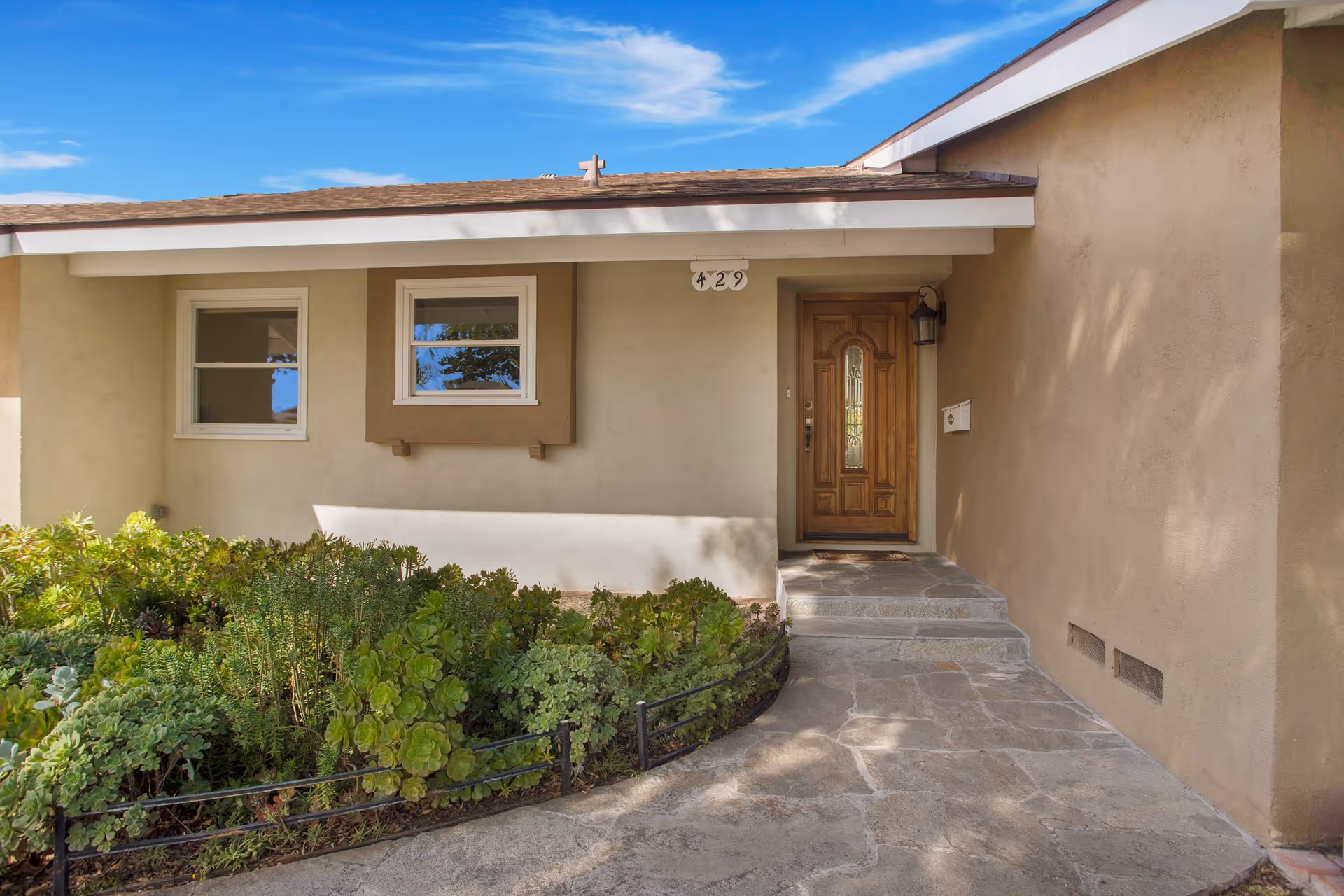 Front entrance of a single-story house with a wooden door, two windows, beige stucco walls, a small garden with green plants, and a stone pathway leading to the door under a clear blue sky.