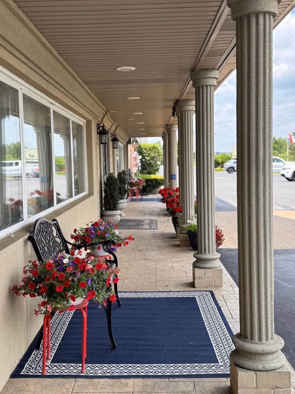 Covered front entrance walkway with fluted columns, benches, and potted red flowers leading toward a parking lot.
