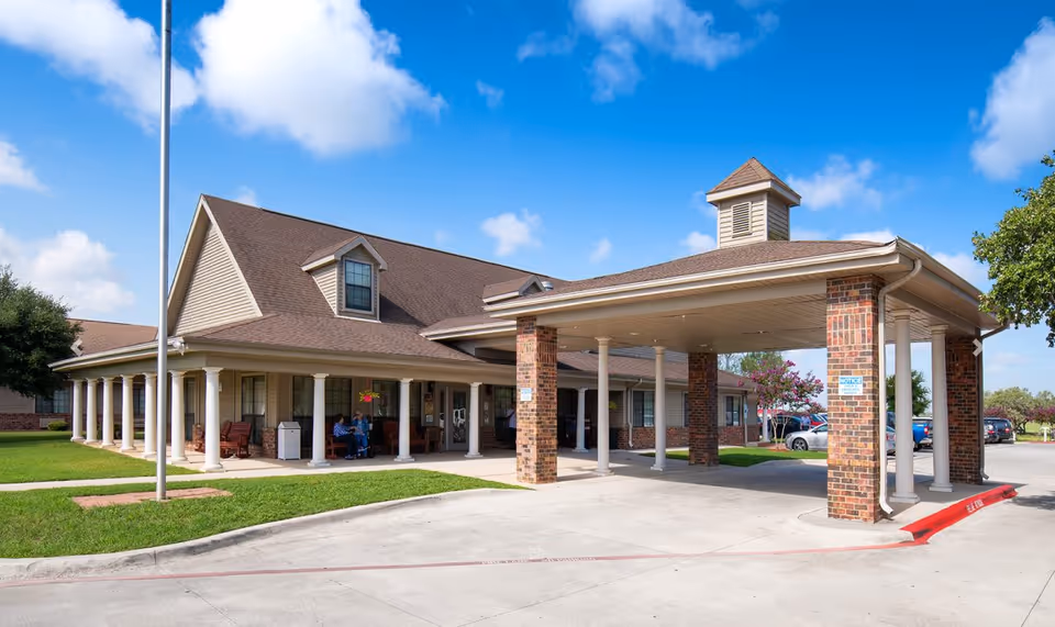Front entrance of a brick-and-siding senior living building with a covered porte-cochère supported by columns under a blue sky.