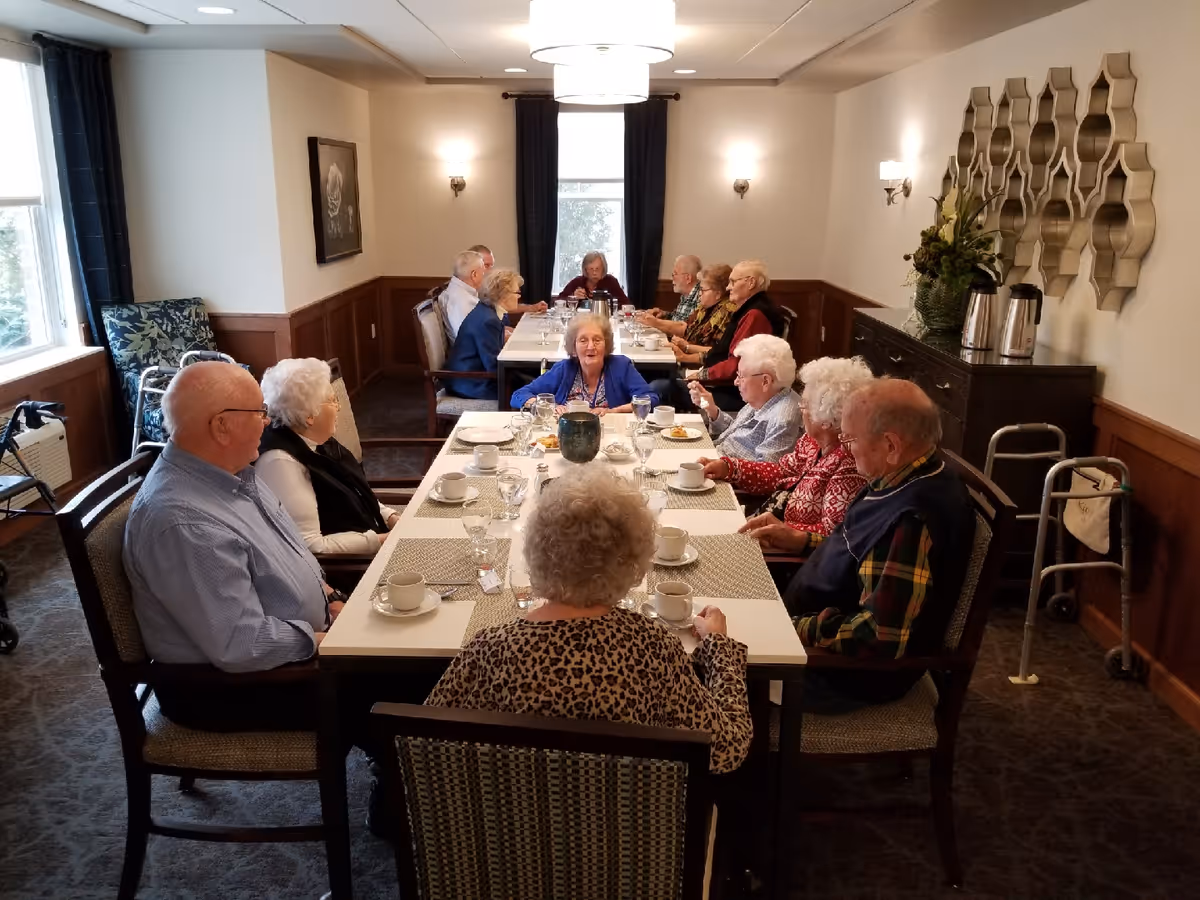 A group of elderly people sitting around a long dining table in a well-lit room, engaged in conversation and having a meal. The room has large windows with dark curtains, wall sconces, a decorative mirror, and a sideboard with coffee pots. Some walkers are visible near the chairs.