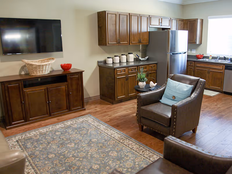 Open-plan living area with leather armchairs, a wall-mounted TV, patterned rug, and a kitchen with wooden cabinets and a stainless refrigerator.