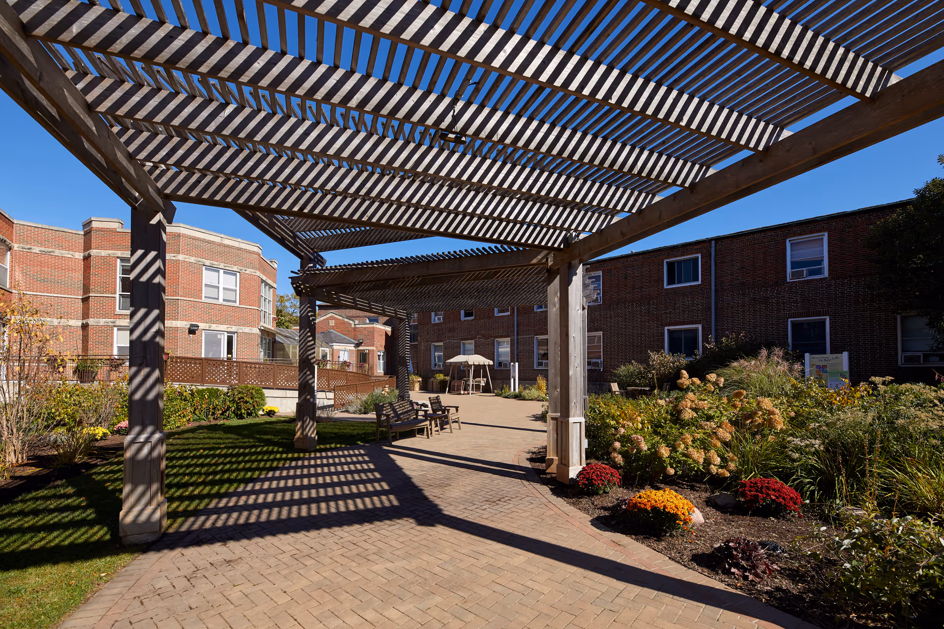 Shaded outdoor courtyard with a wooden pergola casting striped shadows over a brick-paved walkway, benches, and landscaped gardens in front of brick buildings.