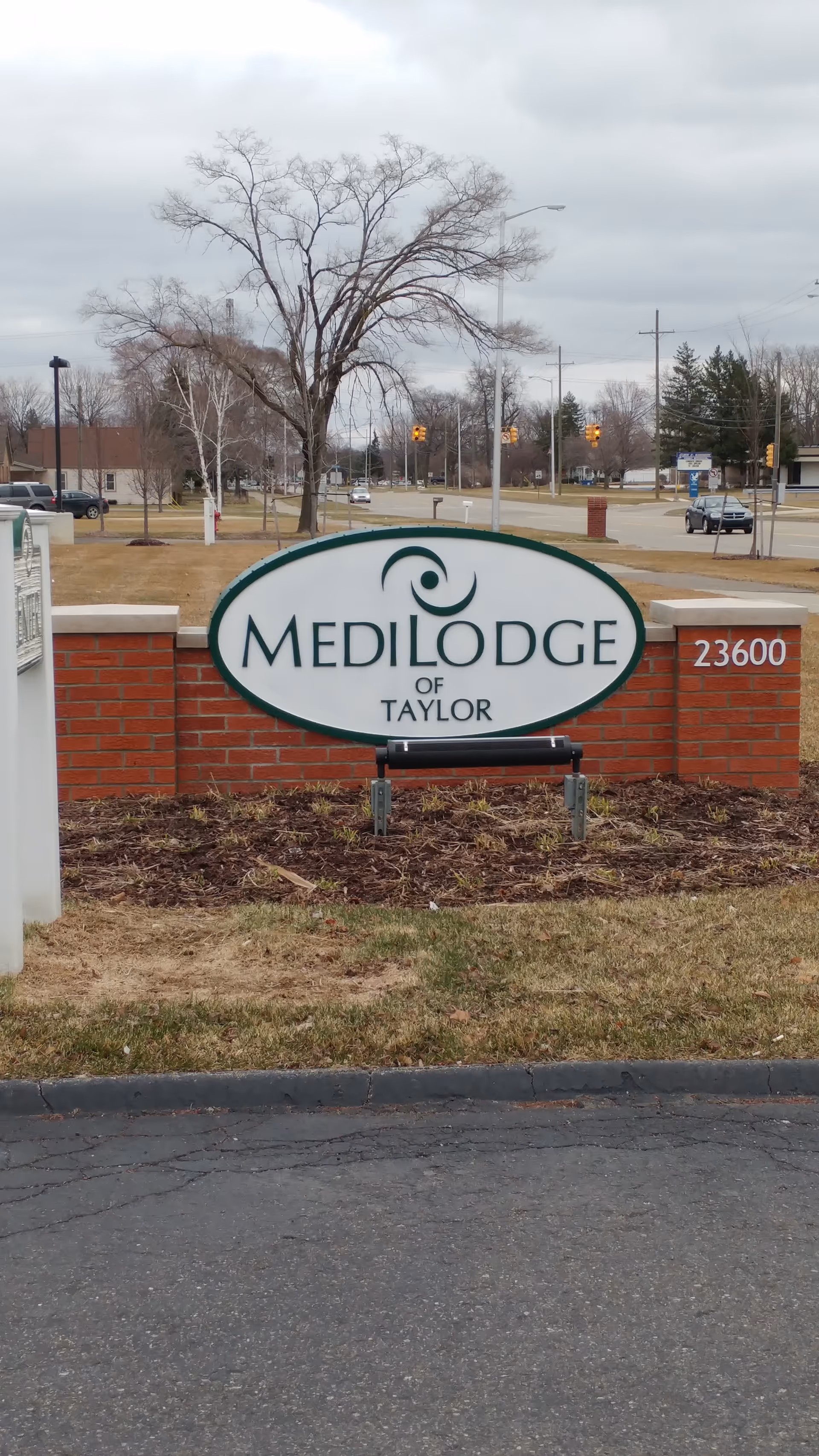 Exterior view of Medilodge of Taylor sign on brick wall with surrounding lawn and road frontage.