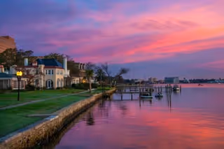 Sunset over a lakeside promenade with boats, grassy lawn, and pastel buildings along water.