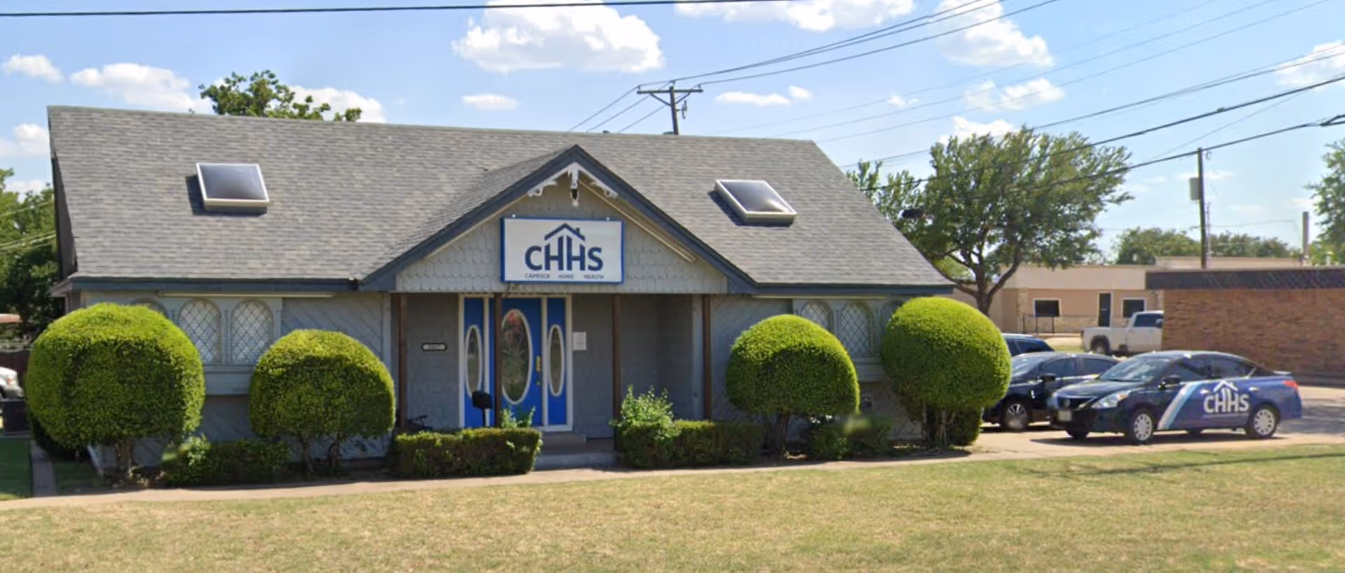 Front view of a small building with CHHS sign, trimmed bushes, and parked cars.