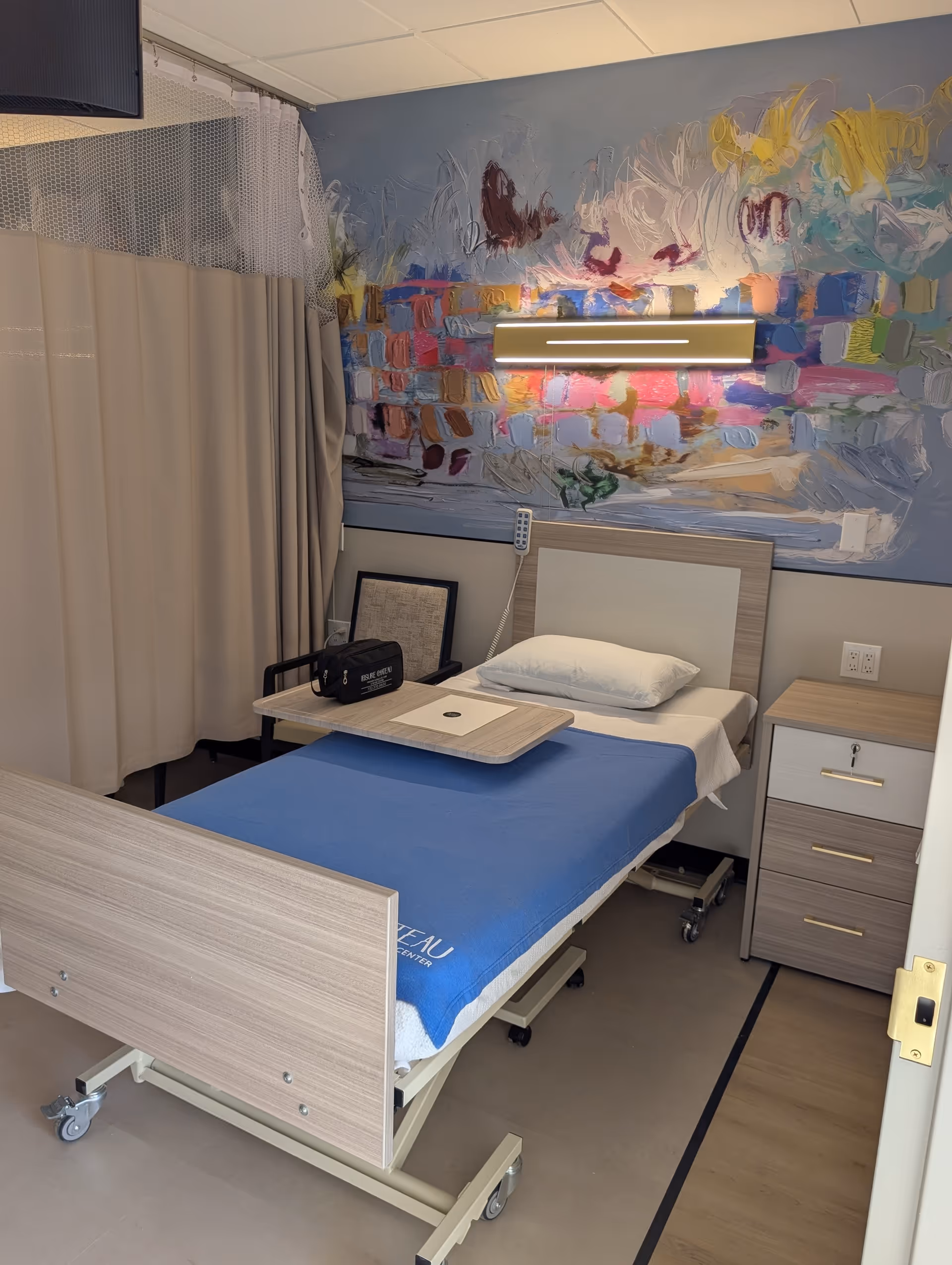 Single hospital-style bed with blue sheet in mural-covered recovery room.