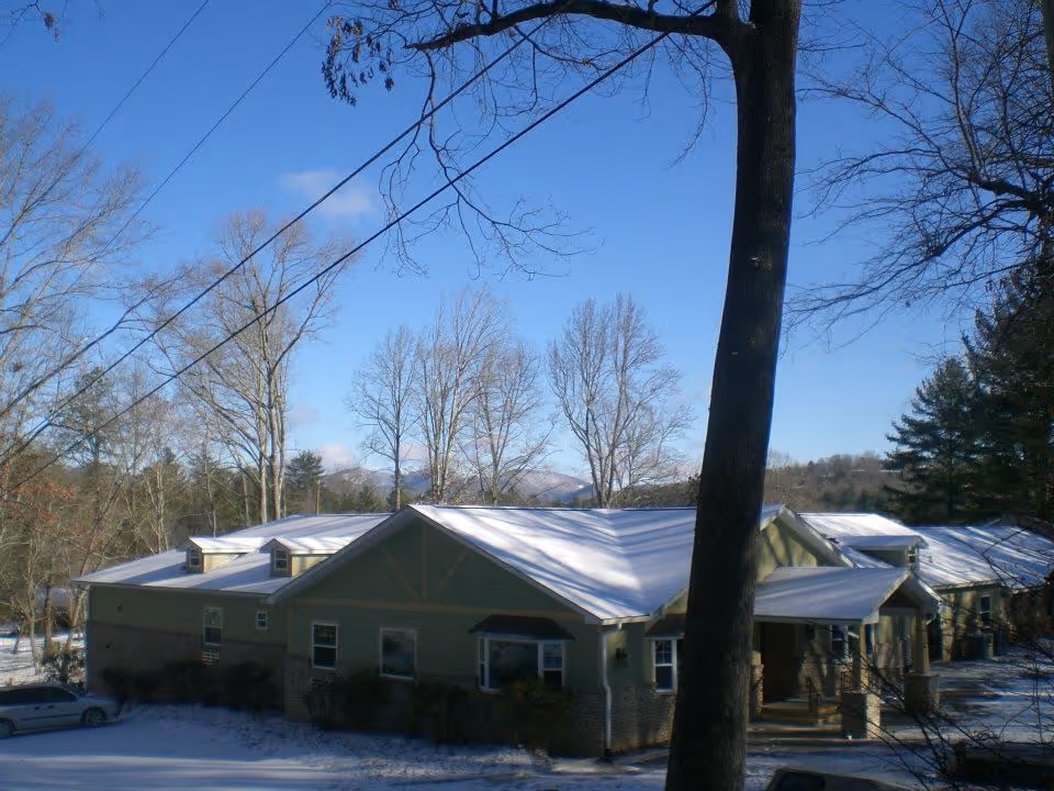 Exterior view of a single-story building with a snow-covered roof and surrounding ground. The building is surrounded by leafless trees under a clear blue sky. A car is parked near the building.