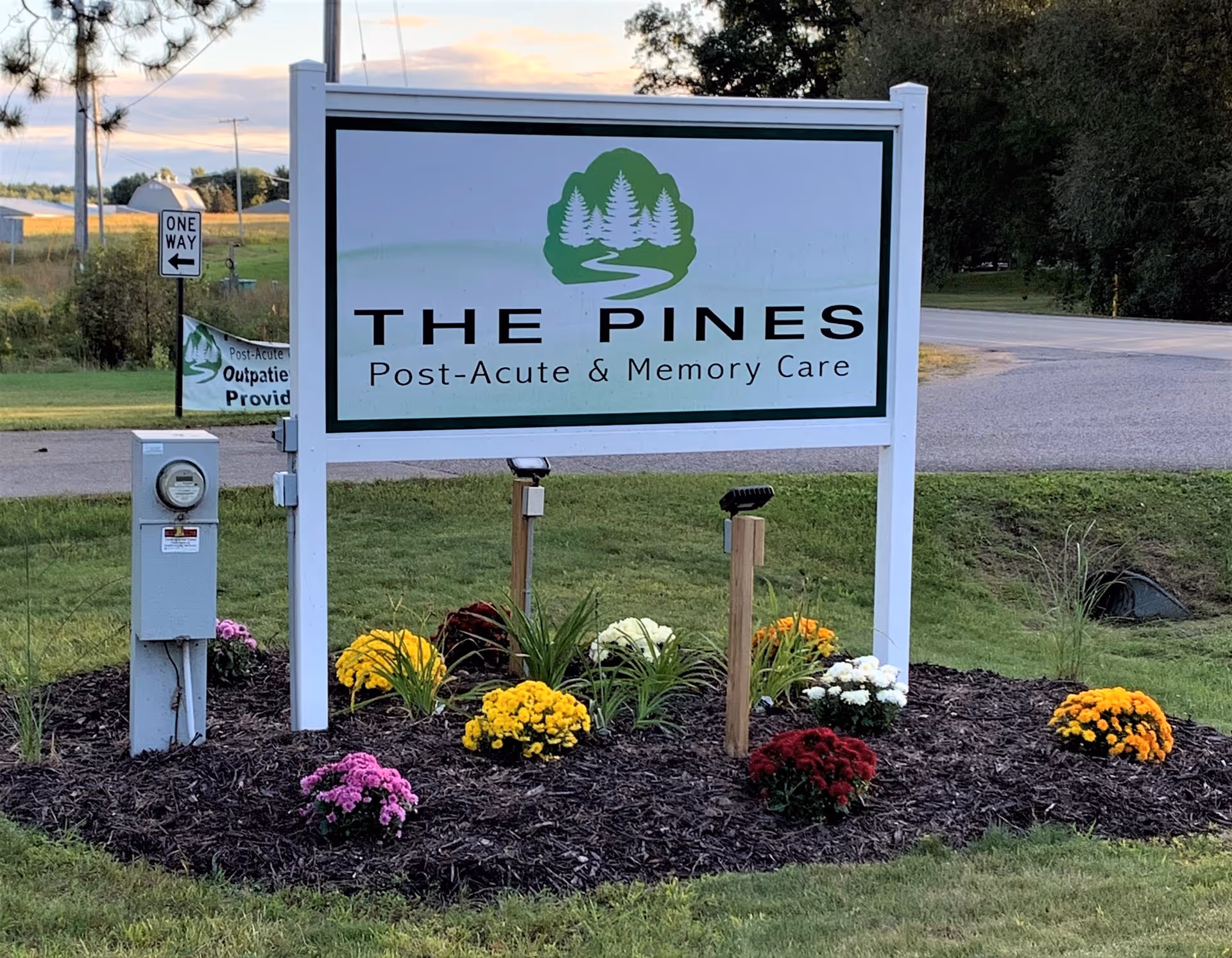 A freestanding sign that reads "THE PINES Post-Acute & Memory Care" surrounded by flowerbeds beside a road.
