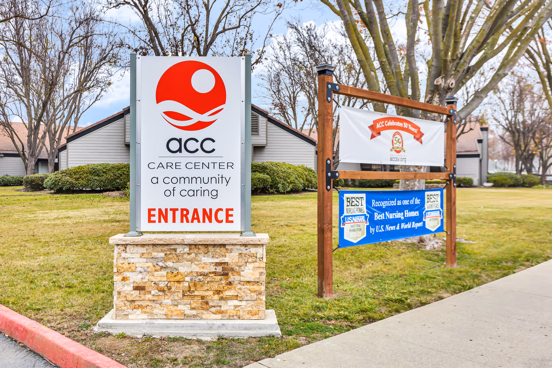 Large ACC Care Center entrance sign on a stone base in front of the facility grounds with banners and buildings in the background.