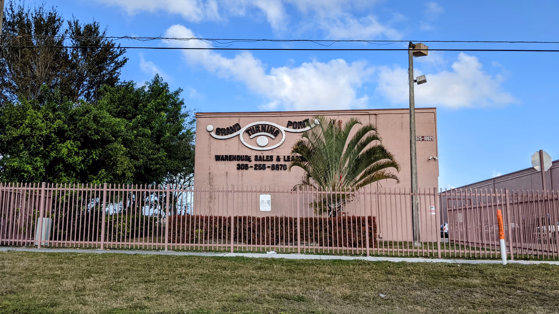 Pink building with a metal fence, palm tree, and Grand Turning Point LLC signage.
