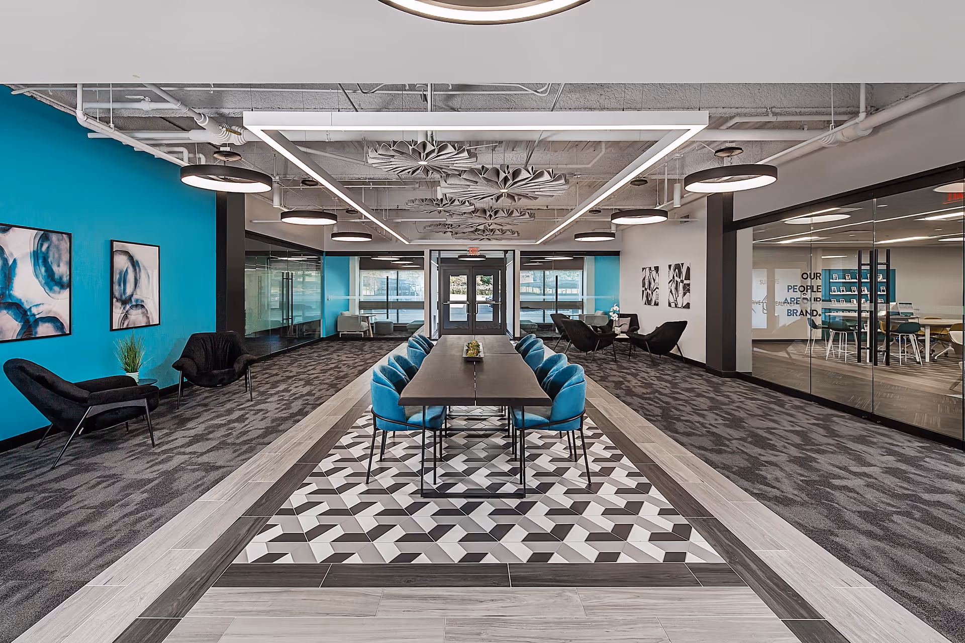 Modern facility lobby with central long table, teal walls, glass offices, geometric floor, and blue chairs.