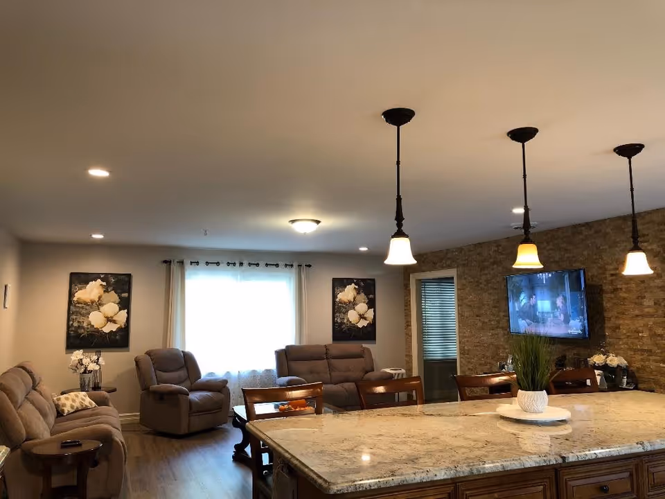 Open living area with recliner sofas, a granite kitchen island with pendant lights, and a wall-mounted TV.