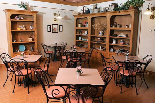 A cozy dining room with several square tables, each surrounded by black wrought iron chairs with pink cushions. The room features wooden flooring, a large wooden cabinet filled with decorative items, and a smaller wooden bookshelf. The walls are adorned with framed pictures and wall-mounted lamps, and small flower arrangements are placed on each table.