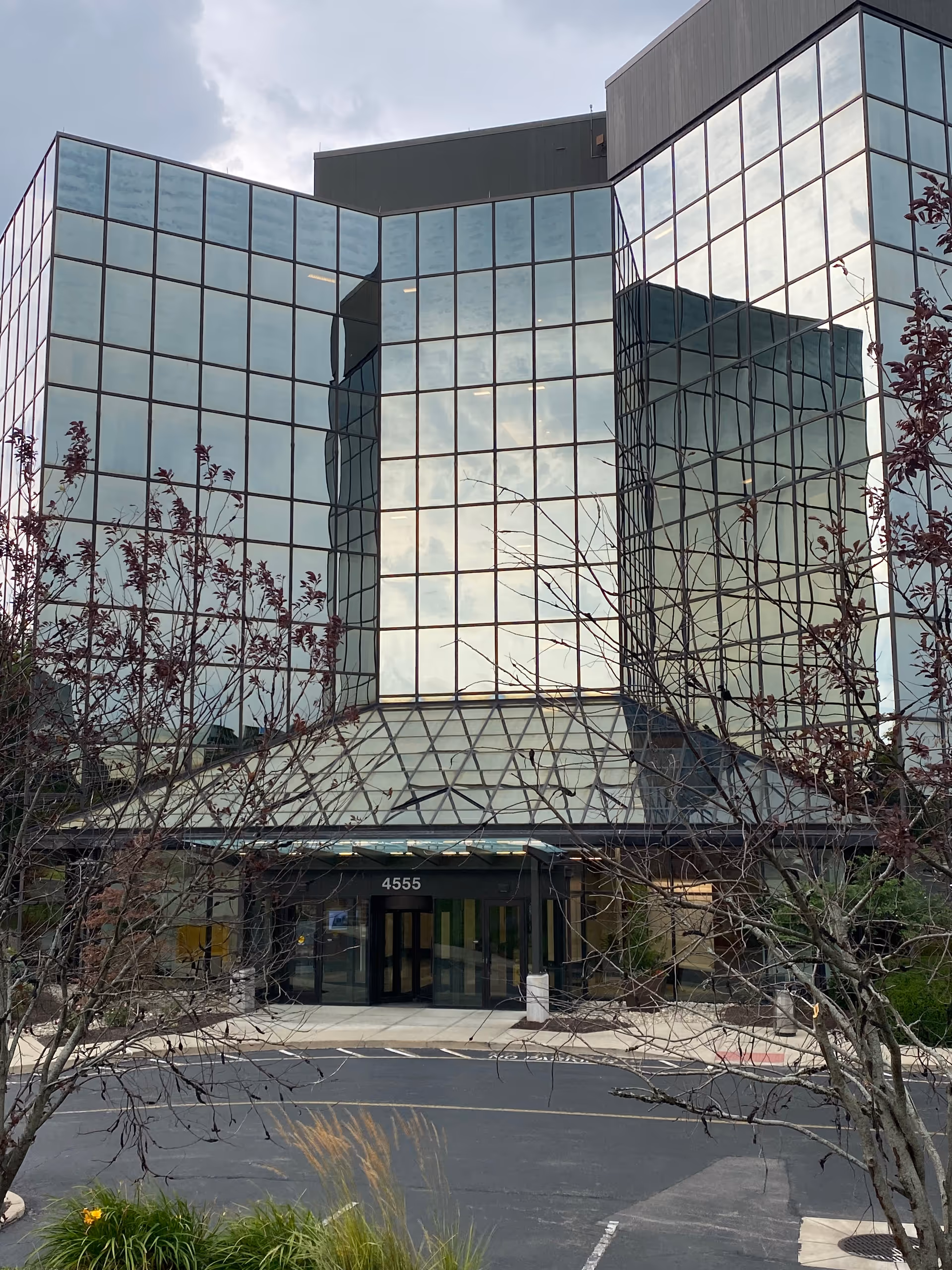 Modern glass-fronted building with a covered entry and bare trees in foreground.