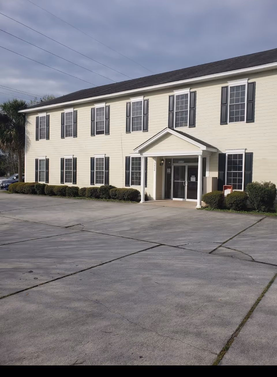 Two-story beige building with black shutters, entrance with columns, and a large concrete parking area.