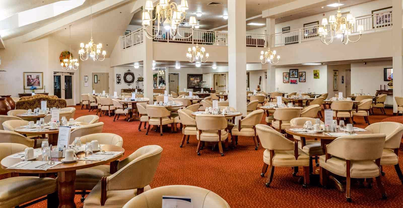 Spacious dining room with multiple round tables set with white cups, glasses, and menus. The room features beige cushioned chairs, red carpet, chandeliers hanging from a high ceiling, and framed artwork on the walls. There is a second-floor balcony overlooking the dining area.