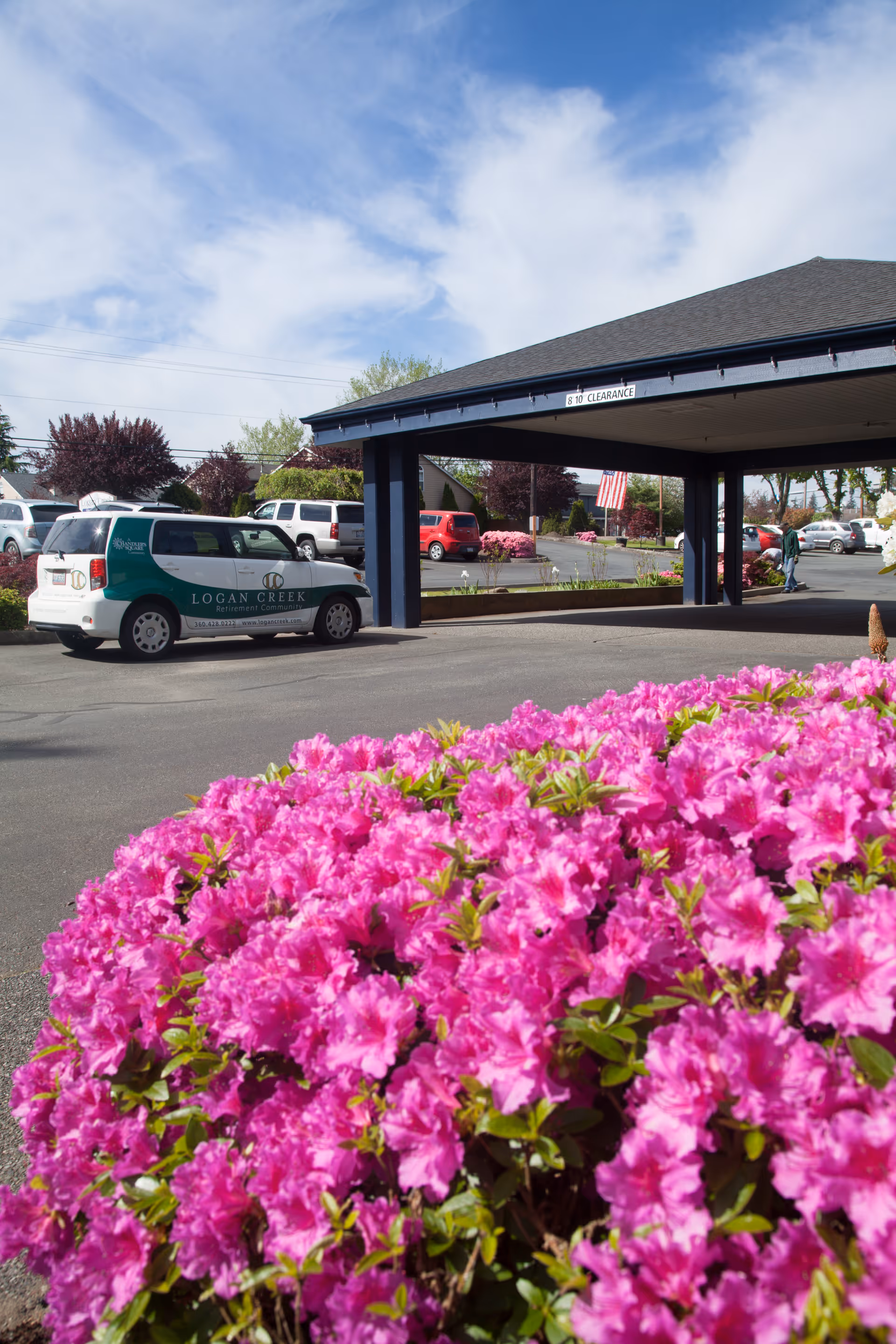 Bright pink flowers in the foreground with a covered driveway and parking lot of Logan Creek Retirement Community in the background, including a Logan Creek branded vehicle and several other parked cars under a partly cloudy sky.