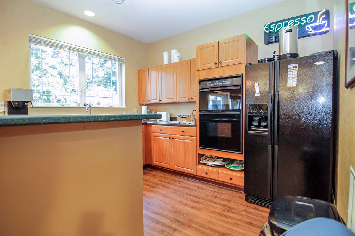 A kitchen area with wooden cabinets, a black double oven, and a black refrigerator. There is a countertop with a coffee maker on it, a window with blinds letting in natural light, and a neon sign above the refrigerator that says 'Espresso' with a coffee cup graphic.