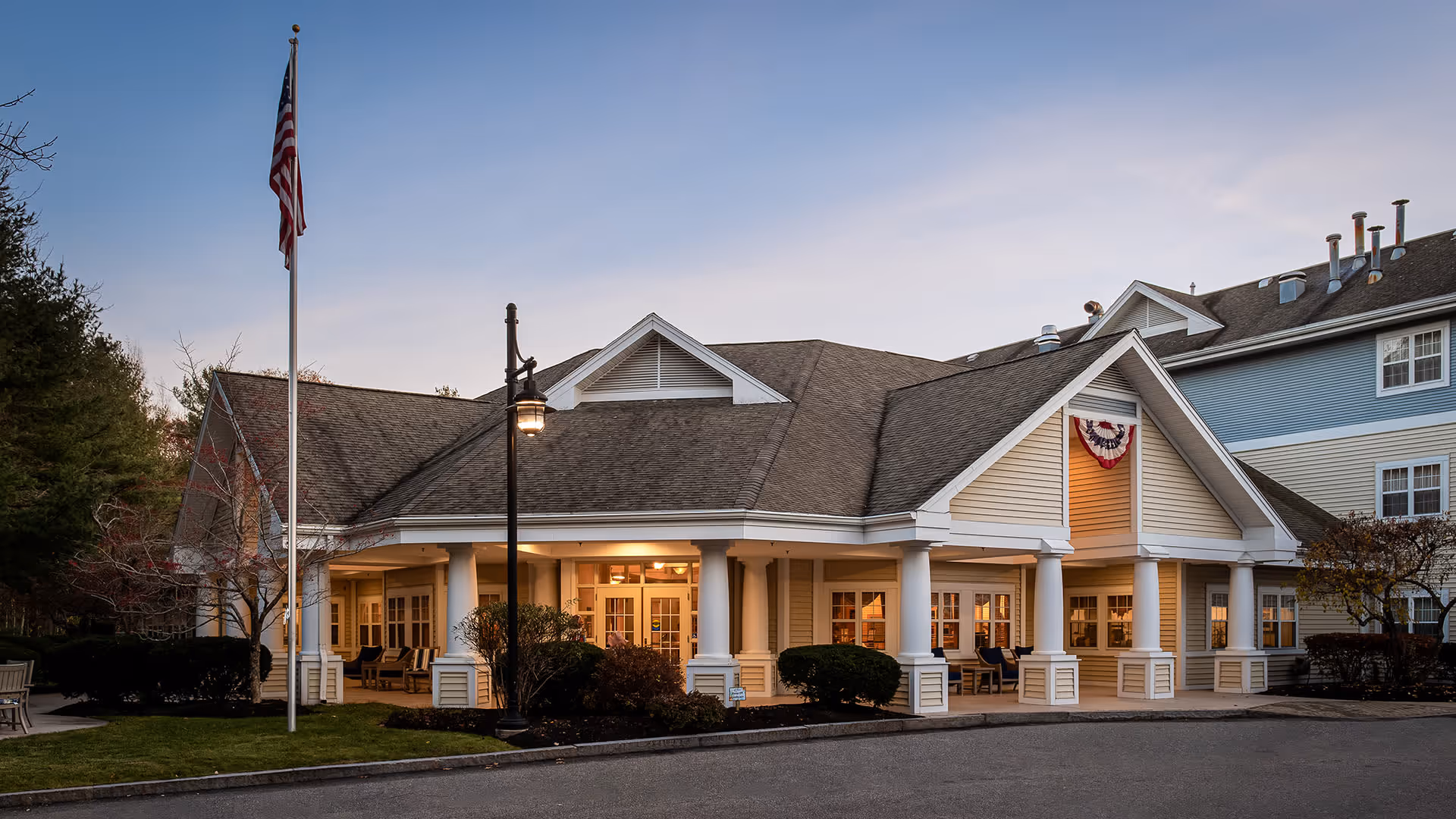 Exterior view of a senior living facility building at dusk with warm lights glowing from inside. The building features a covered porch supported by white columns, an American flag on a flagpole, and a decorative red, white, and blue bunting hanging near the roof peak. There are bushes and trees around the building and a paved driveway in front.