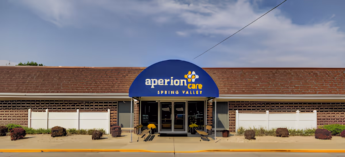 Front entrance of a single-story senior care building with a blue awning reading 'aperion care spring valley' over glass doors and a sidewalk with planters.