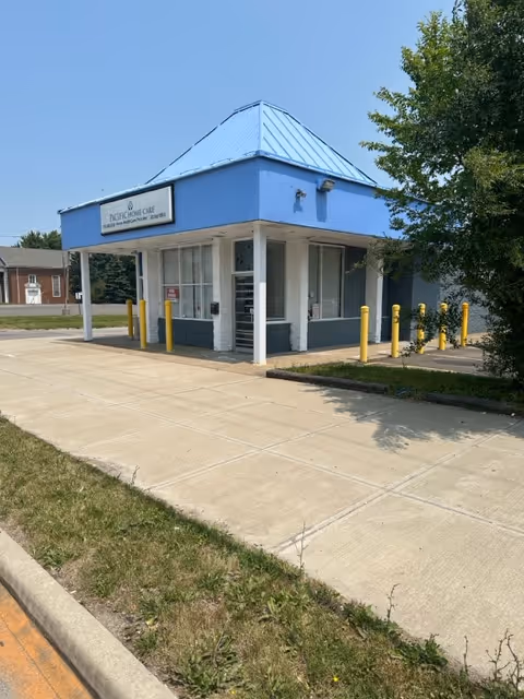 Front exterior of a blue-roofed building with an entry and yellow bollards.