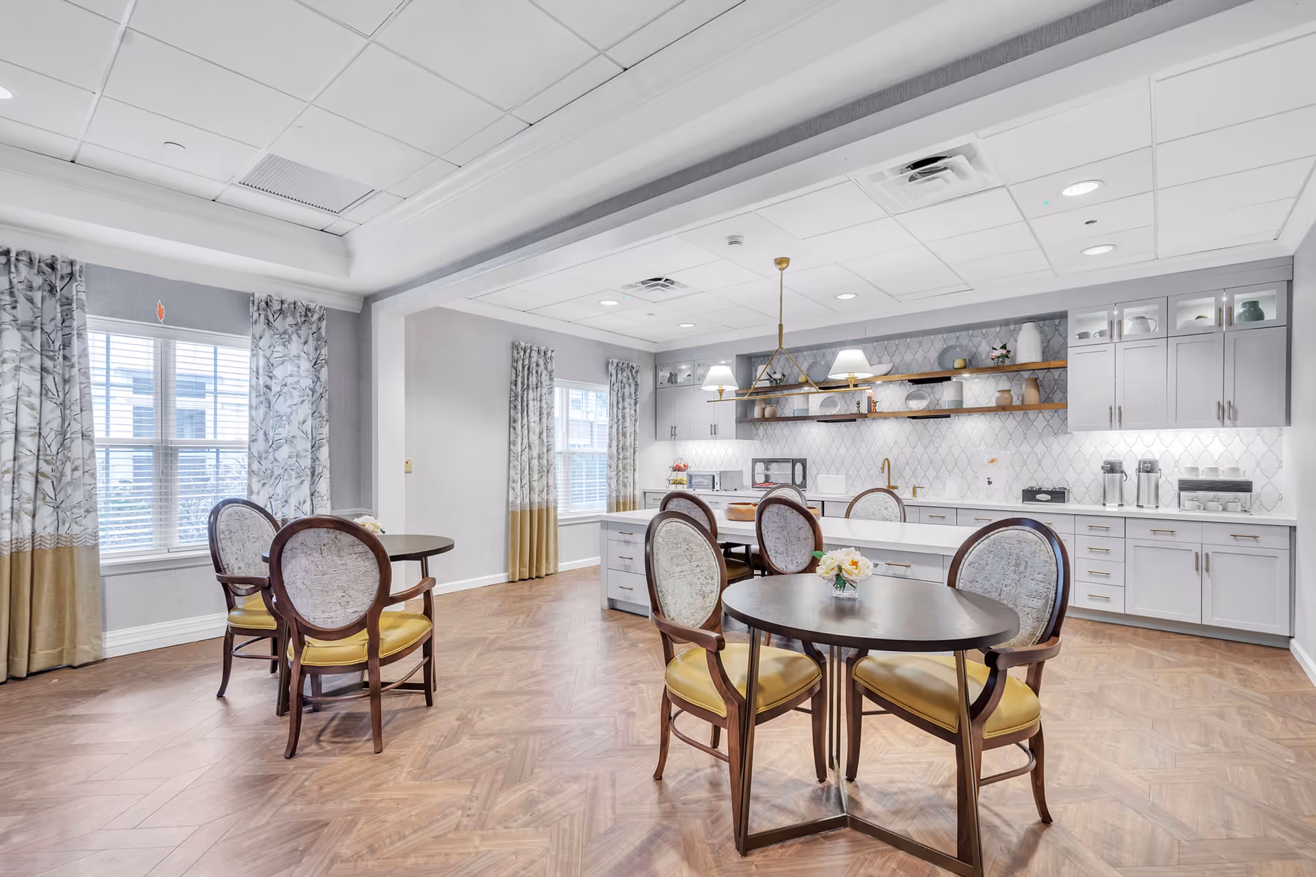 Bright dining area with round tables, upholstered chairs, and light cabinetry in a senior living facility.