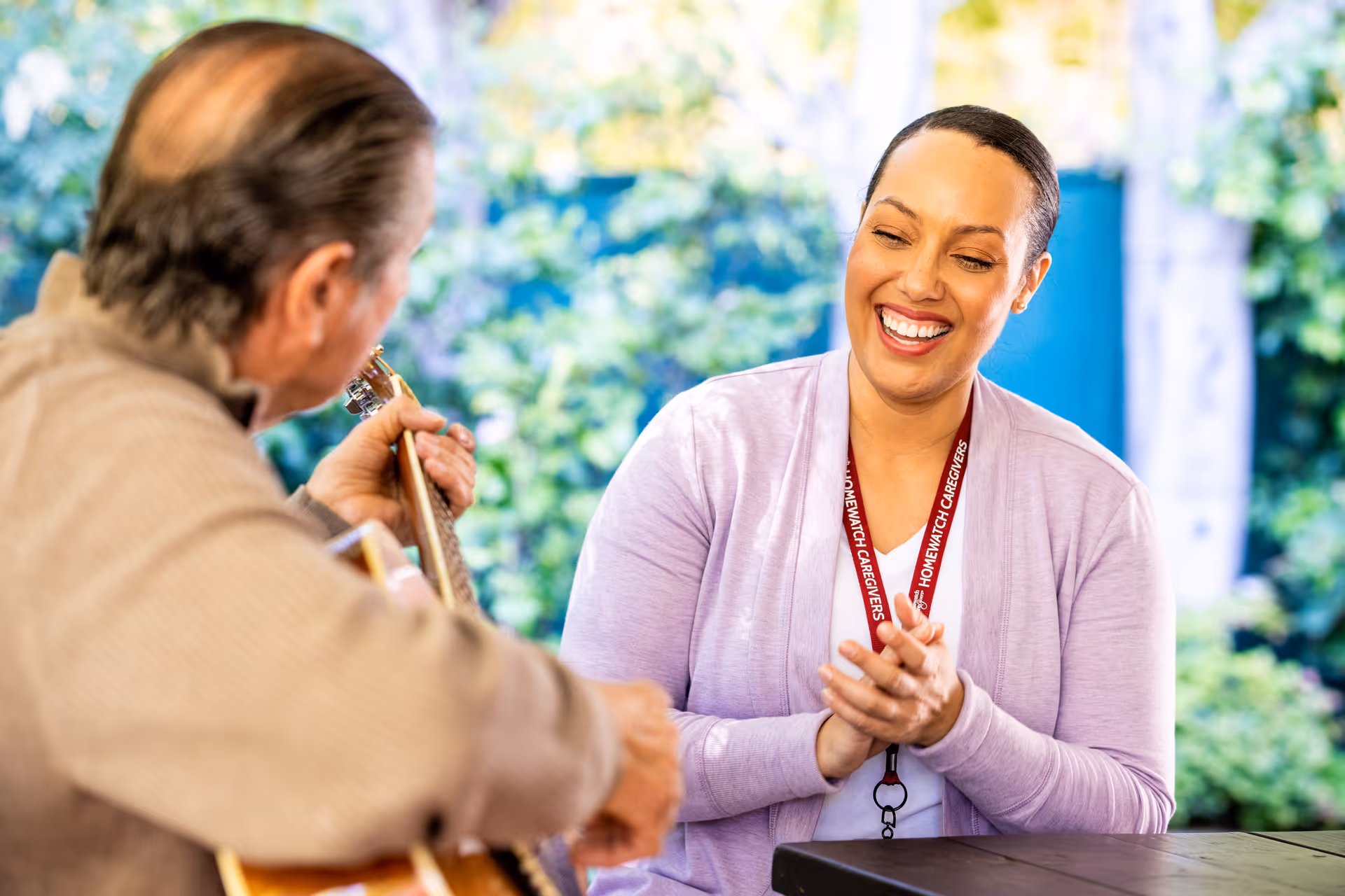 Caregiver and resident share a guitar moment outdoors, smiling in a sunny setting.