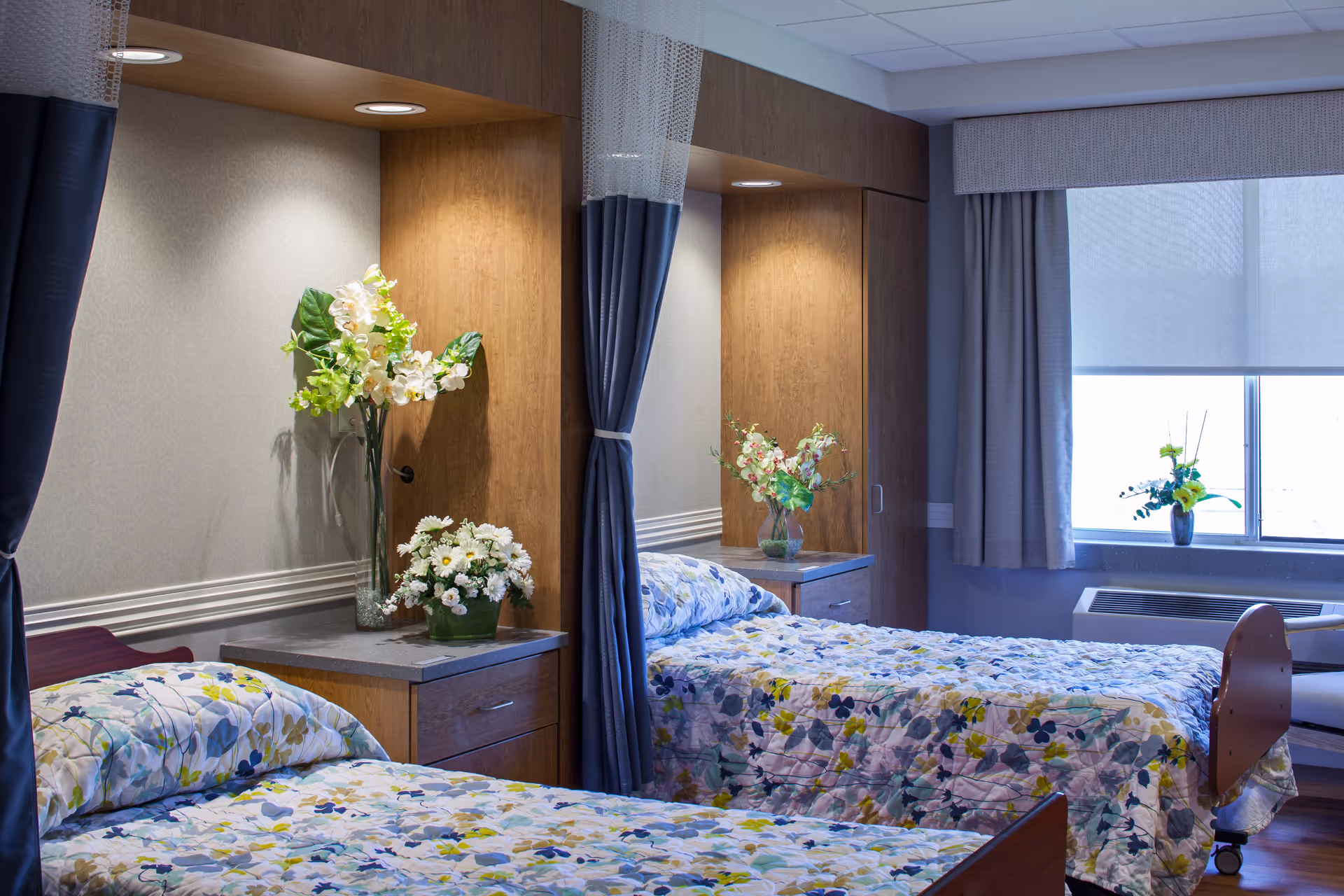 A well-lit bedroom with two single beds, each covered with floral-patterned bedspreads. Between the beds is a wooden nightstand with two vases of flowers. The beds have privacy curtains that can be drawn around them. A window with closed curtains and a small table with a flower vase are visible in the background.
