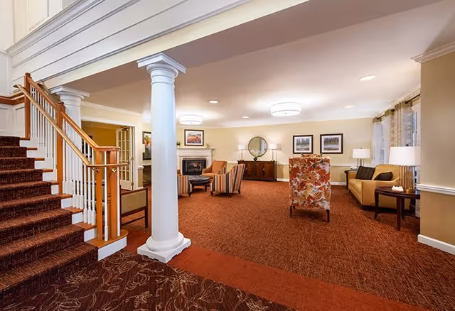 A spacious senior living facility common area with a staircase on the left, a white column in the center, and a seating area with armchairs and a sofa arranged around a fireplace. The room features warm lighting, framed artwork on the walls, and large windows with curtains.