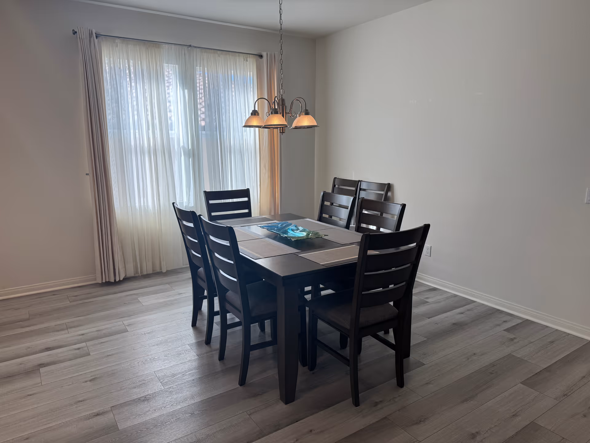 A dining room with a rectangular dark wooden table surrounded by six matching chairs. The table has placemats and a decorative centerpiece. A window with sheer white curtains and beige drapes allows natural light to fill the room. A hanging light fixture with five lamps is positioned above the table. The floor is covered with light-colored wood planks, and the walls are plain white.