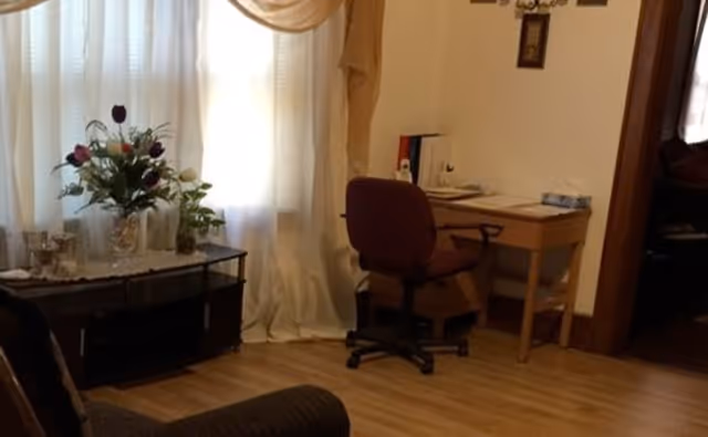A cozy living room corner with a desk, chair, sofa, and floral arrangement near curtains.