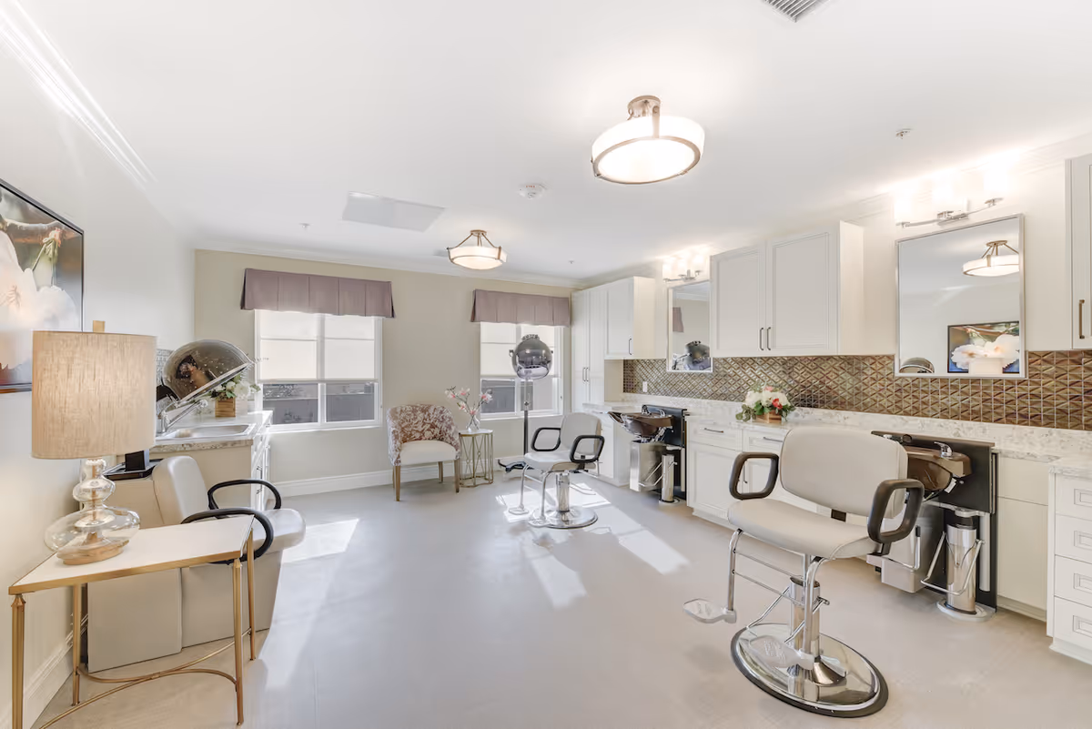 A bright and spacious hair salon room with two salon chairs in front of mirrors and sinks, a hair dryer, a comfortable armchair near two windows with valances, a side table with a lamp, and white cabinetry with a decorative backsplash.