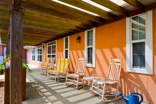 Covered outdoor patio with wooden rocking chairs and lounge chairs lined up against an orange building wall under a wooden pergola.