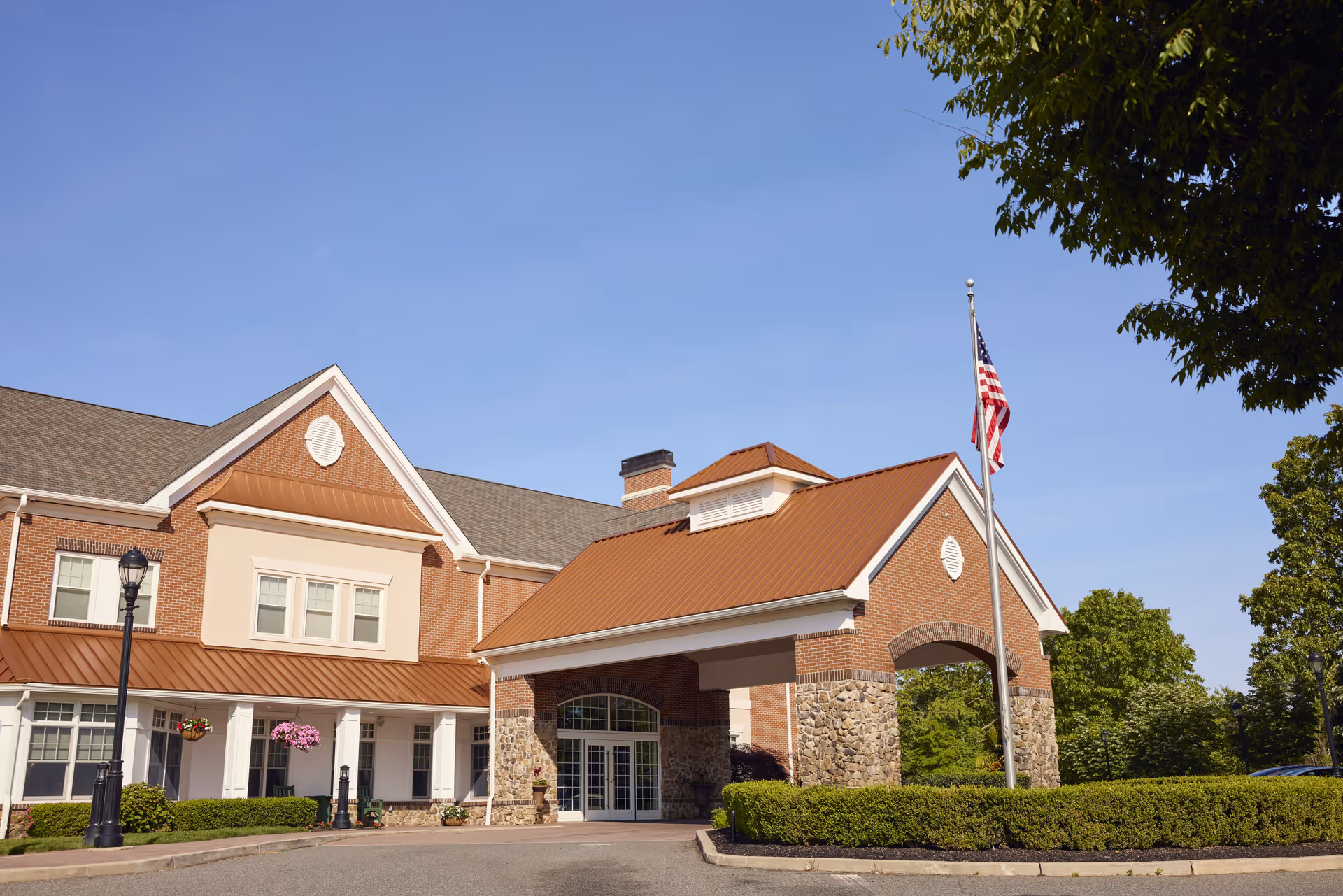 Front exterior view of a senior living facility building with a covered entrance supported by stone pillars, an American flag on a flagpole, and well-maintained landscaping under a clear blue sky.