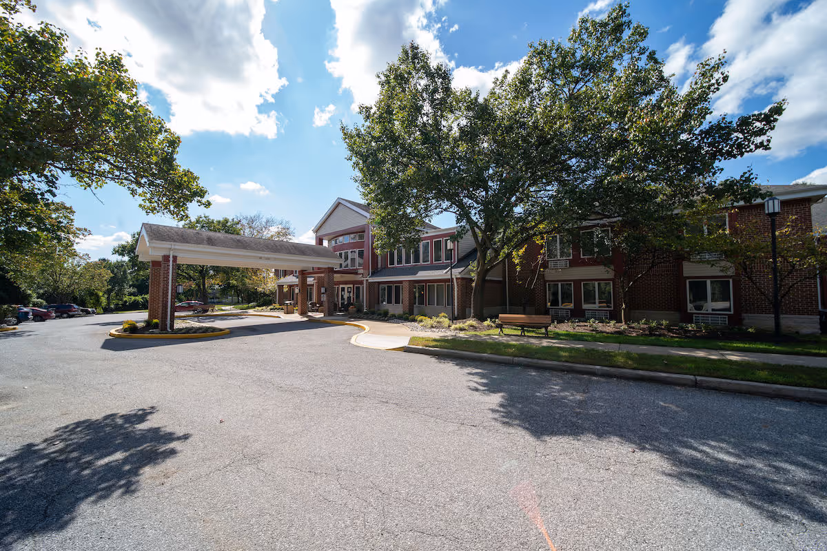 Exterior view of Independence Court of Hyattsville, showing a two-story brick building with a covered entrance driveway, surrounded by trees and a parking area under a partly cloudy sky.