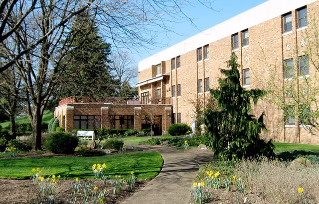 Exterior view of a multi-story brick building surrounded by a landscaped garden with a paved walkway, green grass, trees, and blooming yellow flowers under a clear sky.