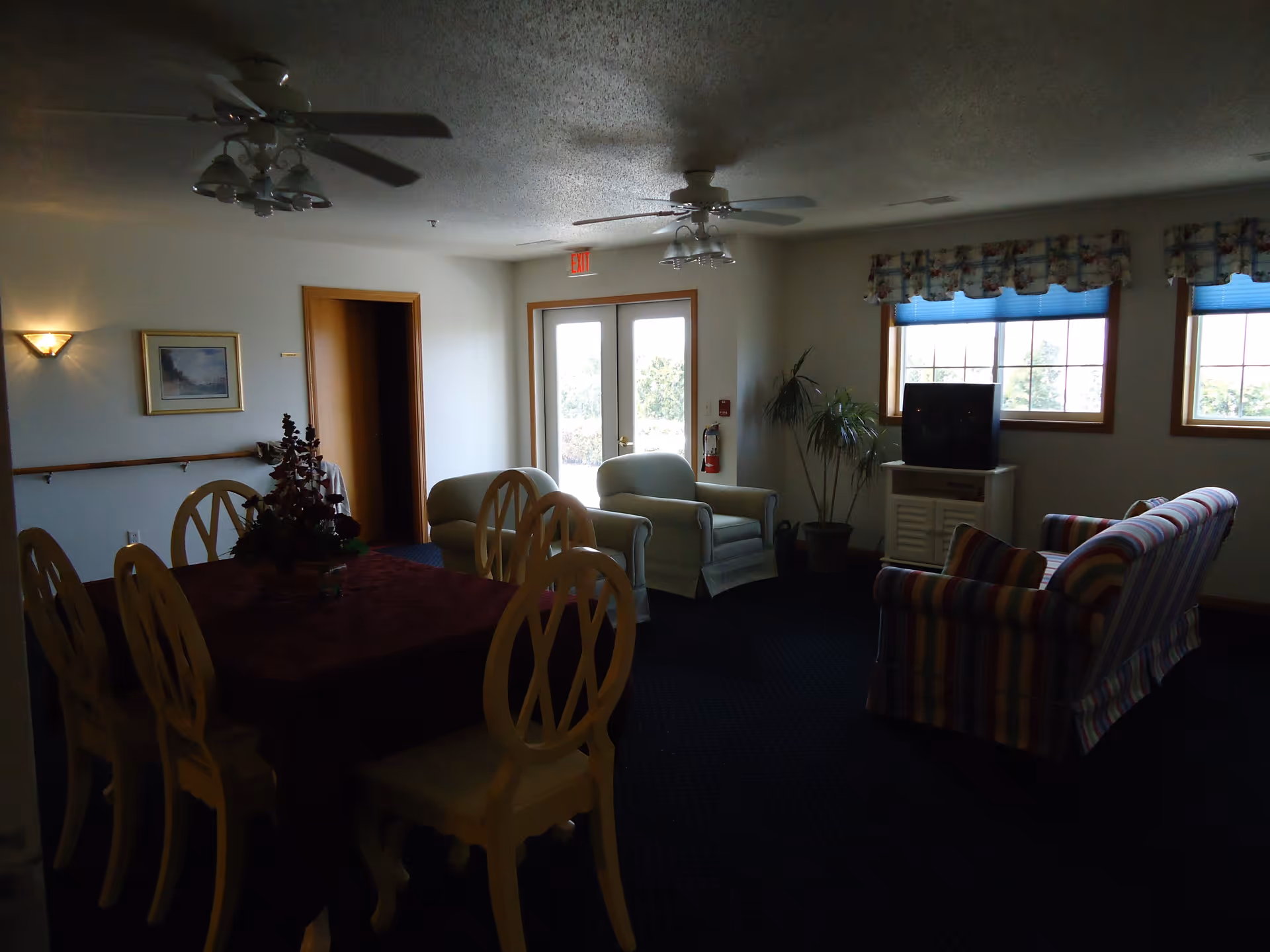 A dimly lit common area in a senior living facility featuring a dining table with six chairs and a floral centerpiece, two armchairs, a striped sofa, a TV on a small stand, two windows with blue blinds and floral valances, a potted plant, and a door with an exit sign above it.
