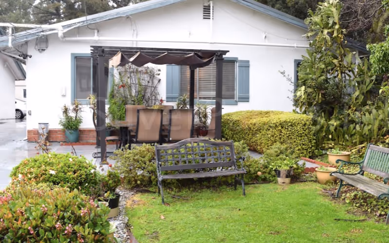 Front yard of a single-story white building with a pergola dining set, benches, and landscaped plants.