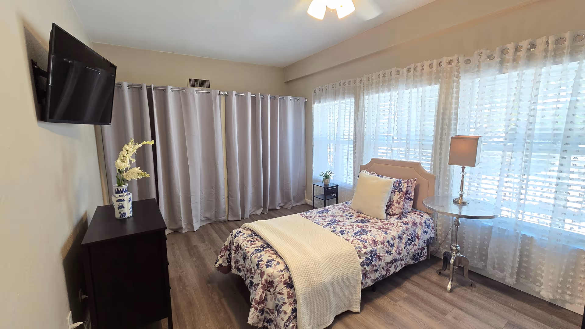 Cozy senior living facility bedroom with floral bedding, bedside table, lamp, and wall-mounted TV.