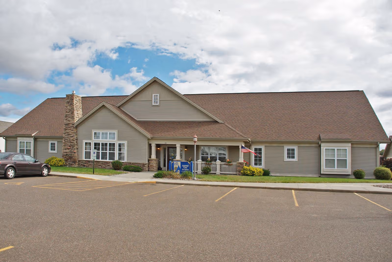 Exterior front view of a single-story senior living facility building with beige siding and a brown roof. There is a stone chimney on the left side, a small porch with white railings and an American flag on the right side. A parking lot with marked spaces is in the foreground under a partly cloudy sky.