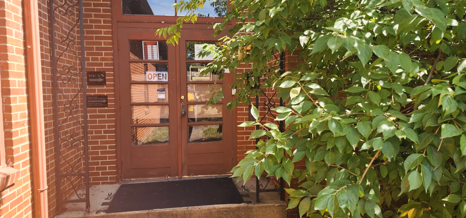 Brick building entrance with brown doors, OPEN sign, and dense greenery.