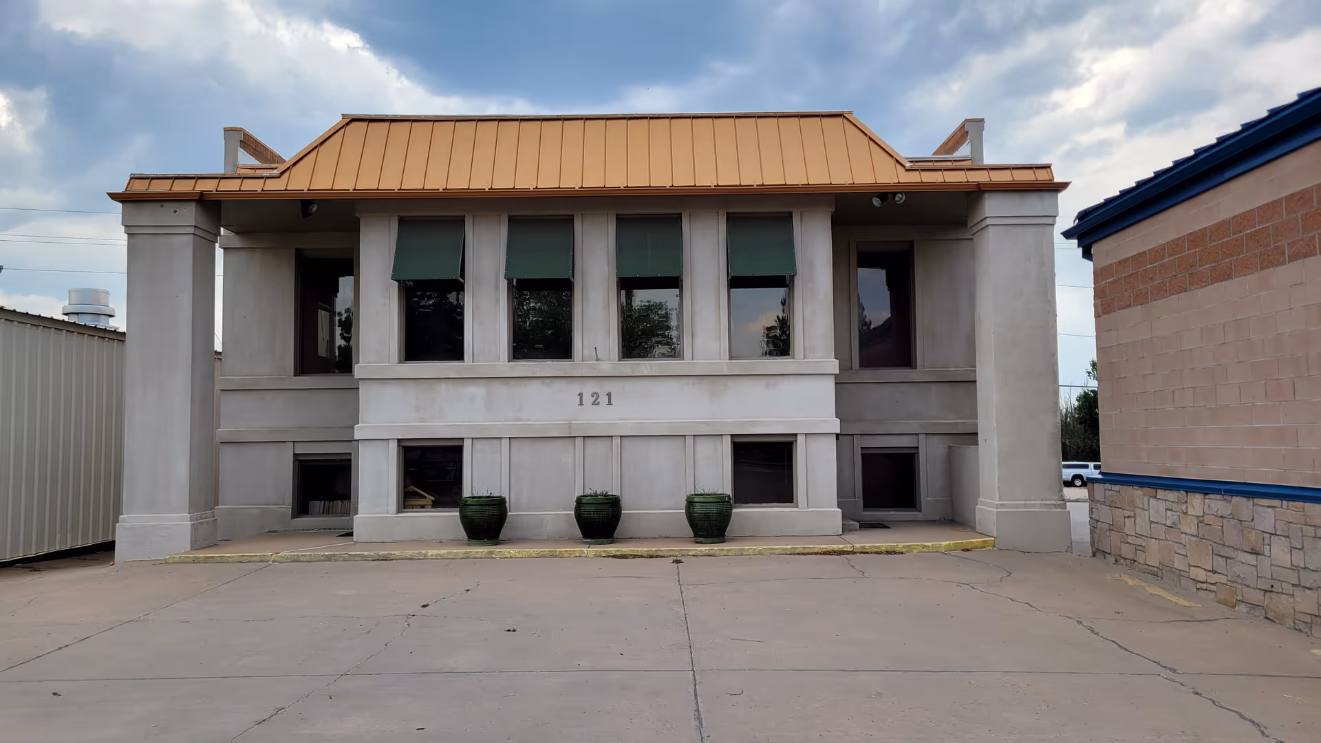 Two-story concrete building with a copper roof and front windows, marked 121, in a parking lot.
