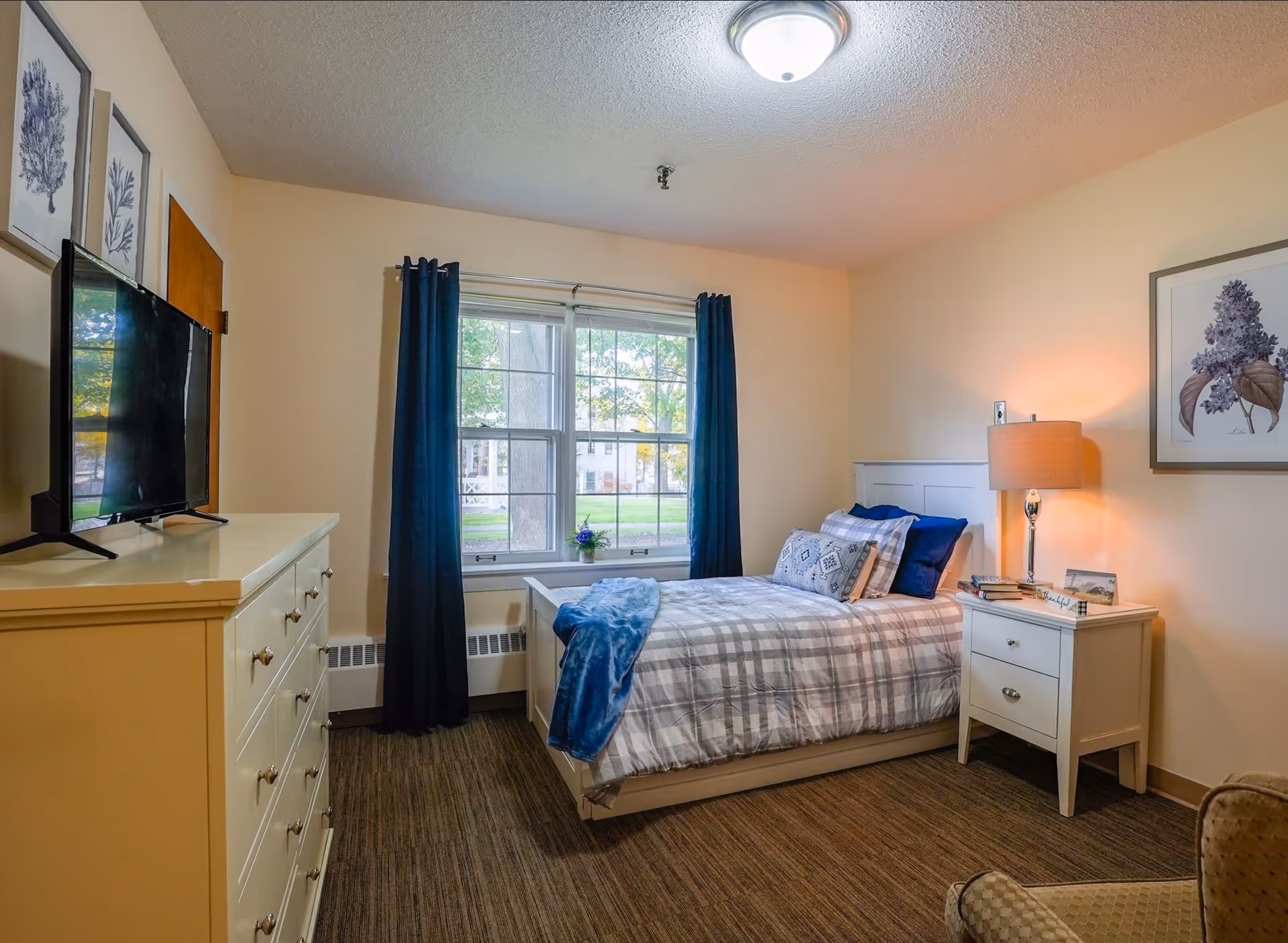 A senior living bedroom with bed, dresser, nightstand, lamp, and window with blue curtains.
