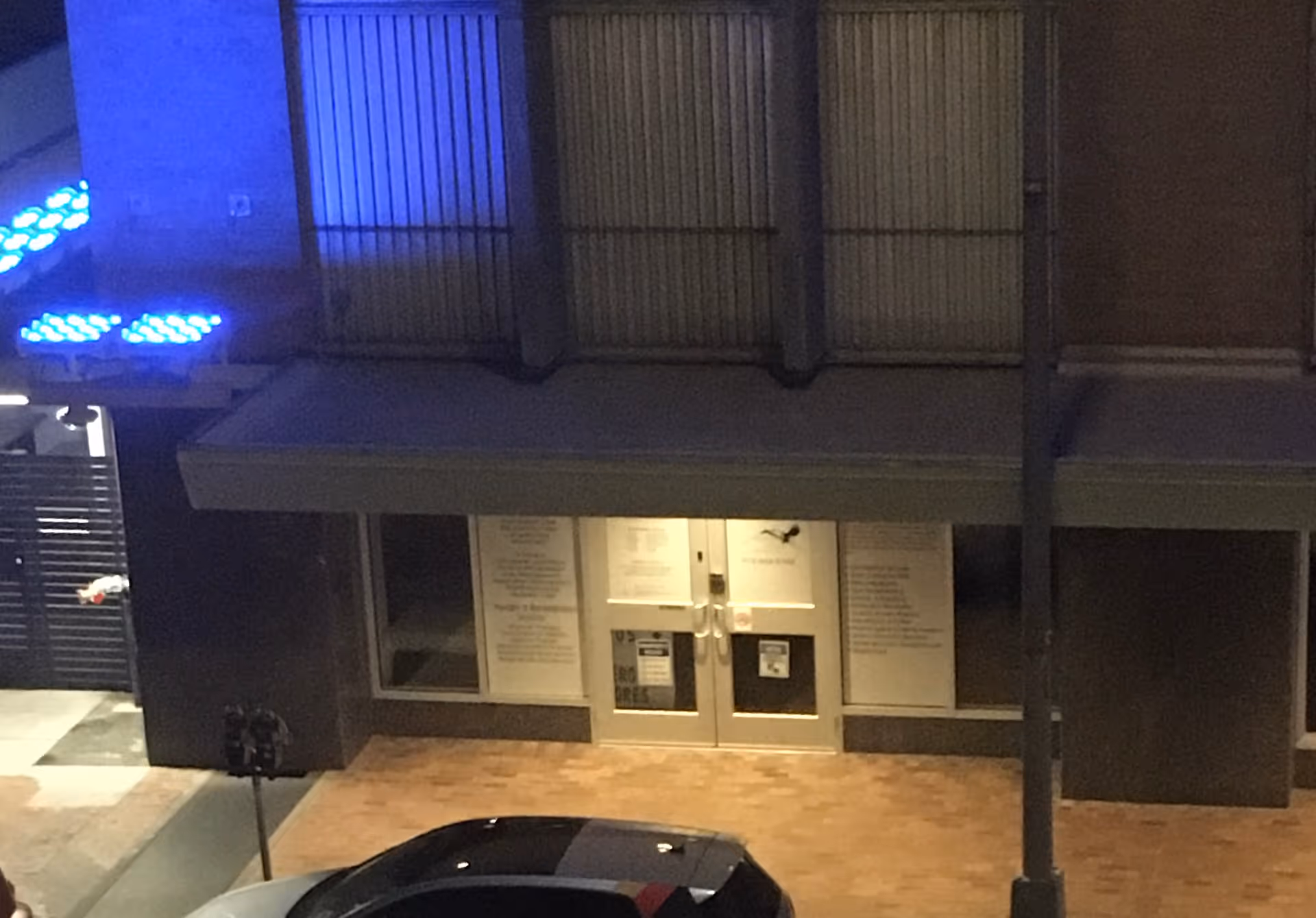 Night exterior of a storefront building with blue-lit accents and glass double doors at the entrance.